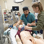 Emergency medicine residents work on a mannikin in a simulation. The mannikin is in a hospital bed in a simulated emergency.