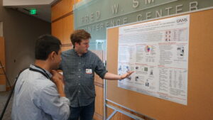 Perry Caviness points to a research poster while another person looks on. Perry is a man with short hair and wearing a button-up shirt. The other person is a man with show dark hair. His back is to the camera.