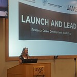 Samantha Kendrick is a woman speaking from behind a podium in an auditorium with a screen behind her saying Launch and Lead: Research Career Development Workshop