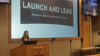 Samantha Kendrick is a woman speaking from behind a podium in an auditorium with a screen behind her saying Launch and Lead: Research Career Development Workshop