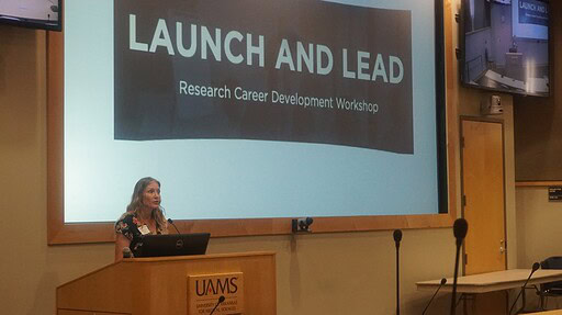 Samantha Kendrick is a woman speaking from behind a podium in an auditorium with a screen behind her saying Launch and Lead: Research Career Development Workshop