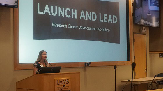 Samantha Kendrick is a woman speaking from behind a podium in an auditorium with a screen behind her saying Launch and Lead: Research Career Development Workshop