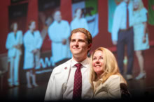 A close-up of a student posing for a photo with a family member onstage with a large live image of faculty in white coats behind them.