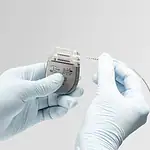 Image shows Onward Medical’s implantable ARCIM System, which is designed to address symptomatic blood pressure instability in individuals with chronic spinal cord injury. Device is small and silver with tubing and is held in gloved hands.
